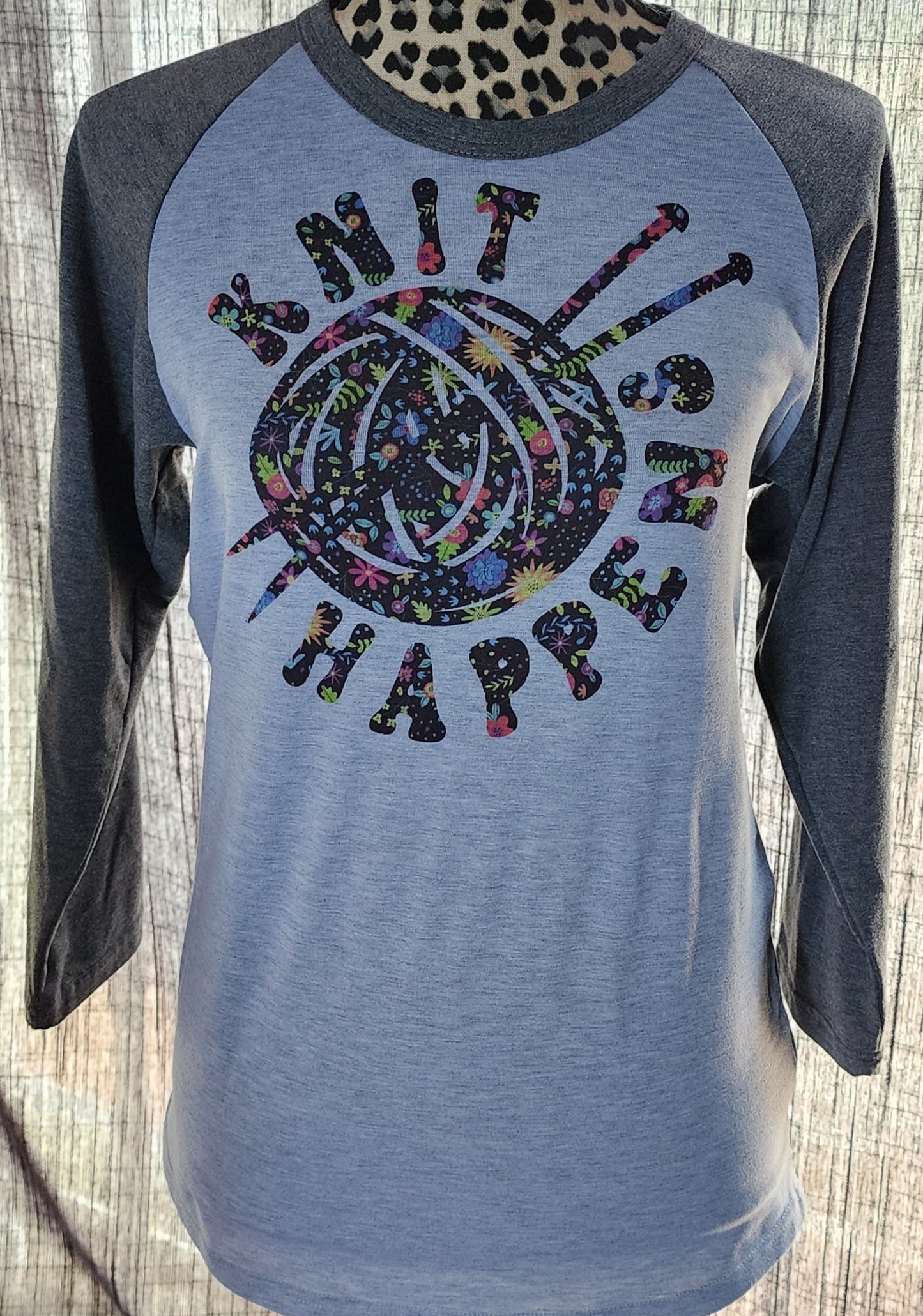 Grey shirt body with dark grey sleeves featuring a design with black ink with various colored floral pictures. The design is the words 'Knit Happens'  wrapped around a ball of yarn with knitting needles. 